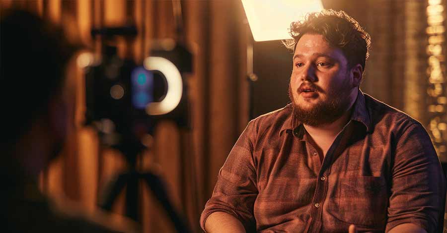 Um homem com barba e cabelo cacheado está sendo entrevistado em um ambiente com iluminação quente. Ele usa uma camisa xadrez de manga comprida e está sentado, falando diretamente para a câmera. Ao fundo, há um tripé com uma câmera e um anel de luz, e cortinas marrons desfocadas. A cena sugere uma gravação de depoimento ou documentário.