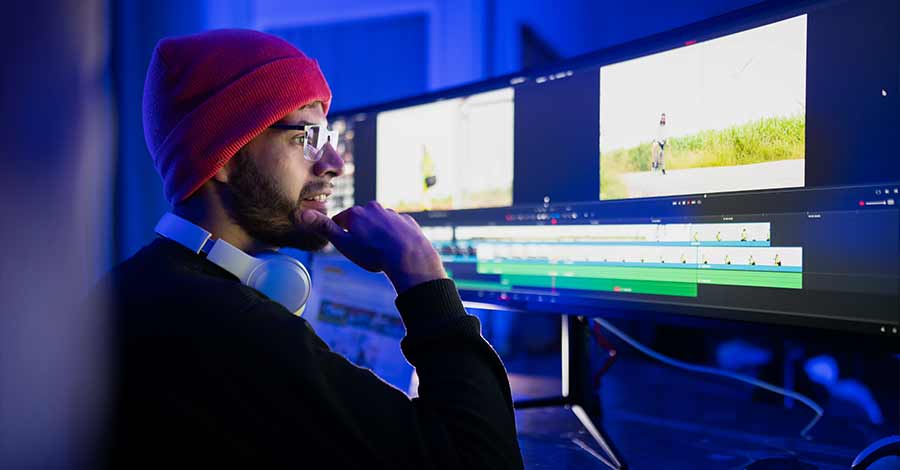 Editor de vídeo com touca vermelha trabalhando em correção de cor para alcançar estética cinematográfica em projeto de filmagem.