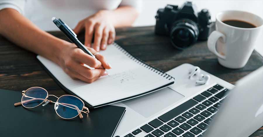 Pessoa escrevendo em um caderno sobre uma mesa com notebook, óculos, câmera fotográfica, fones de ouvido e uma xícara de café, sugerindo um ambiente de trabalho criativo ou de produção de conteúdo.