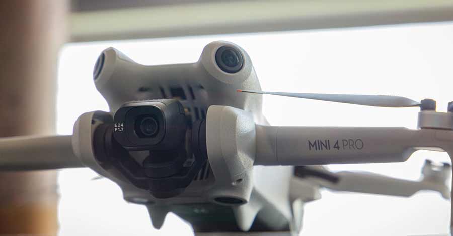 Close-up frontal de um drone DJI Mini 4 Pro, mostrando a câmera principal com especificações na lente (E24 F1.7) e sensores ao redor. As hélices do drone estão parcialmente visíveis, e o nome do modelo está escrito no braço lateral.