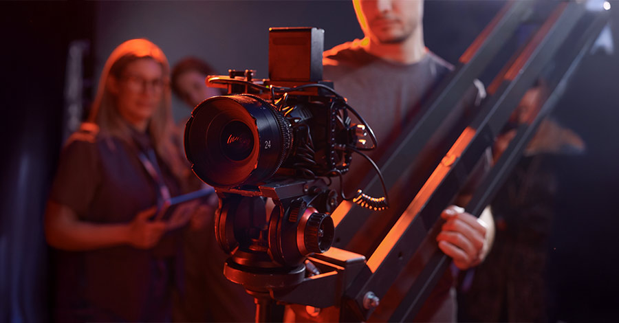 Câmera profissional de cinema montada em grua, com profissionais audiovisuais ao fundo em ambiente de estúdio com iluminação quente.