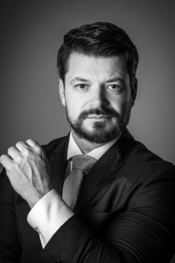 Homem de terno preto posa com expressão séria, olhando para a câmera, com a mão apoiada no punho, em fundo escuro. Foto em preto e branco com iluminação dramática