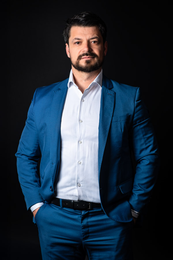 Homem de terno com leve sorriso e postura confiante, encarando a câmera em fundo escuro. Expressão equilibrada entre seriedade e acessibilidade.