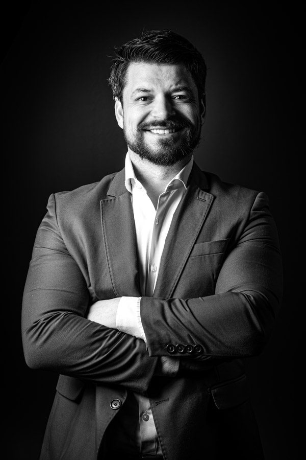 Retrato em preto e branco de um homem sorridente com barba e cabelo curto, usando paletó e camisa social clara, de braços cruzados e postura confiante, sobre fundo escuro.