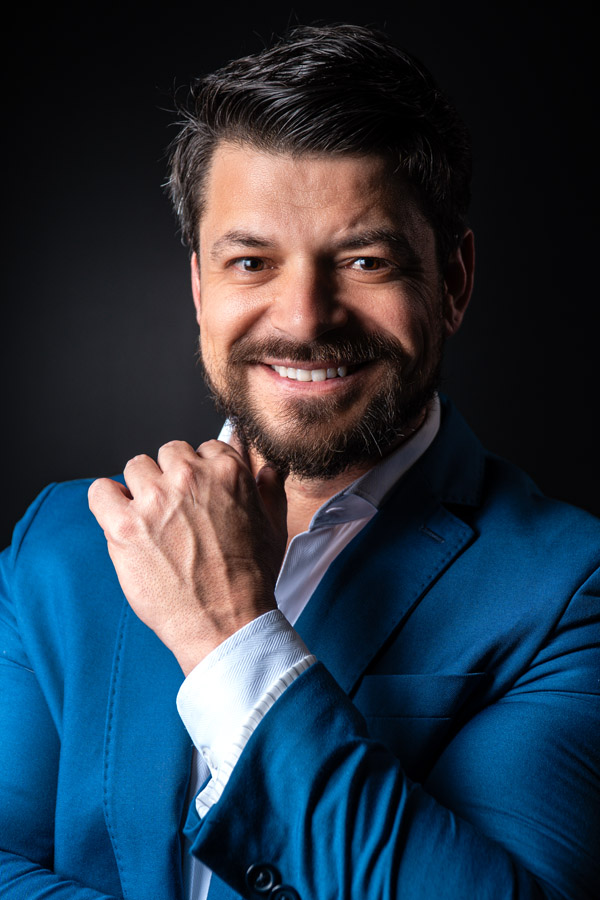 Retrato colorido de um homem sorridente com barba e cabelo escuro, vestindo terno azul e camisa branca, com a mão apoiada no queixo em um gesto descontraído, sobre fundo preto.