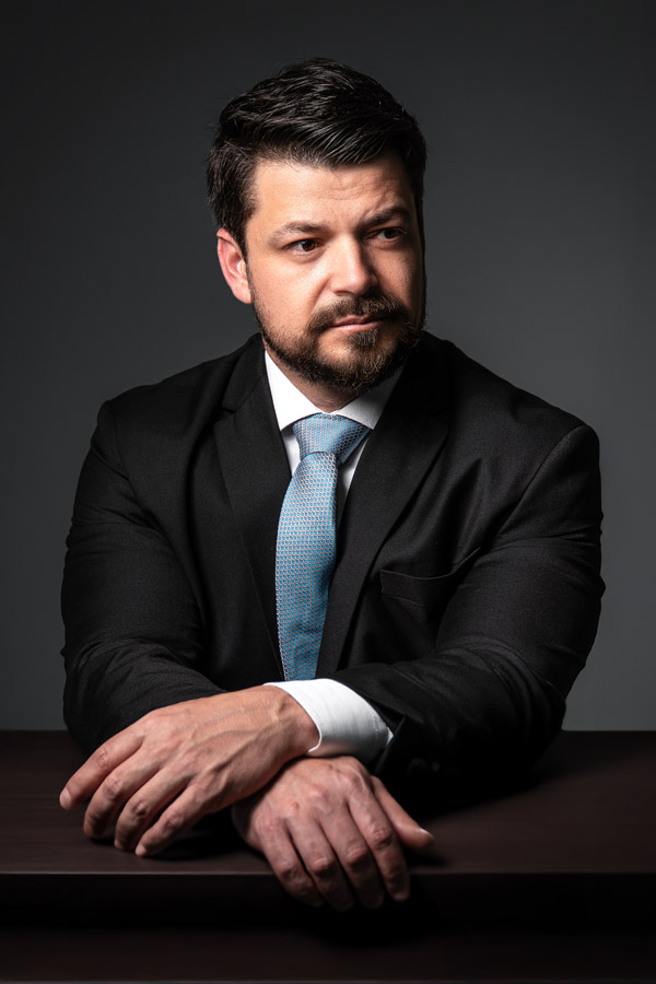 Homem de terno observa à distância com expressão pensativa, levemente virado para a esquerda, em fundo escuro. A luz destaca o rosto e transmite introspecção.