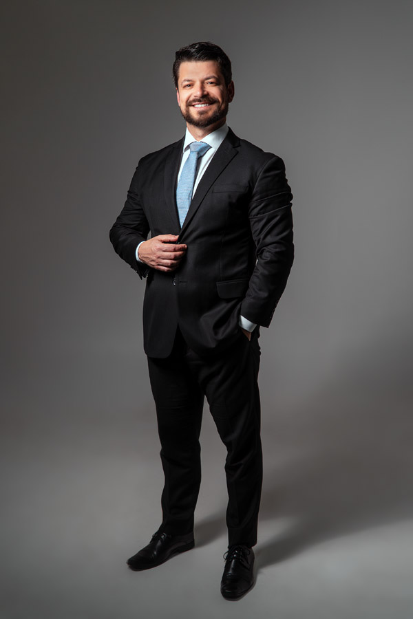 Homem de negócios em pé, sorrindo, vestindo terno preto com gravata azul clara e camisa branca. Ele está posando de forma confiante com uma mão no bolso e a outra ajustando o paletó. O fundo é neutro e profissional, destacando a formalidade do ensaio corporativo.