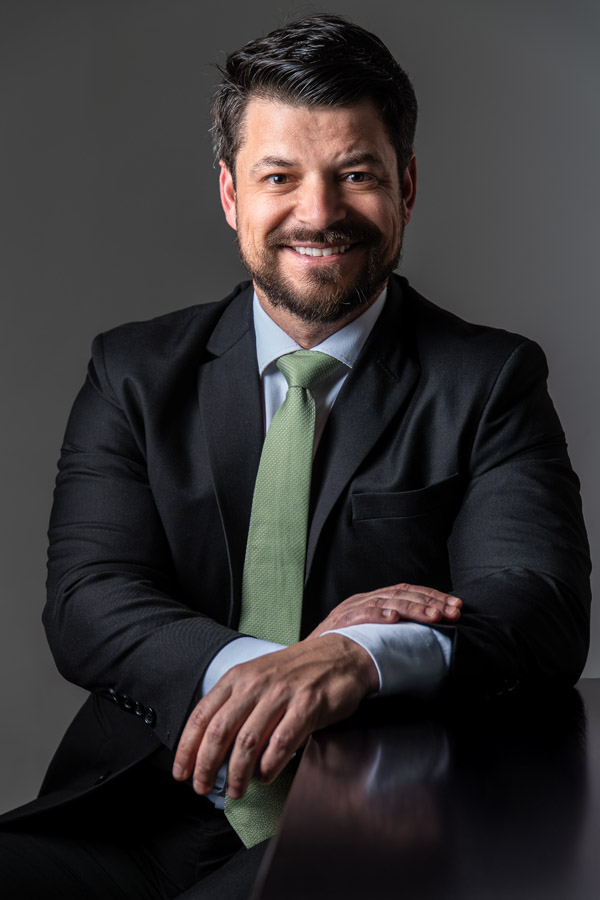 Homem sorrindo de forma leve e simpática, usando terno, com fundo escuro e iluminação suave. Expressão acessível e postura amigável.