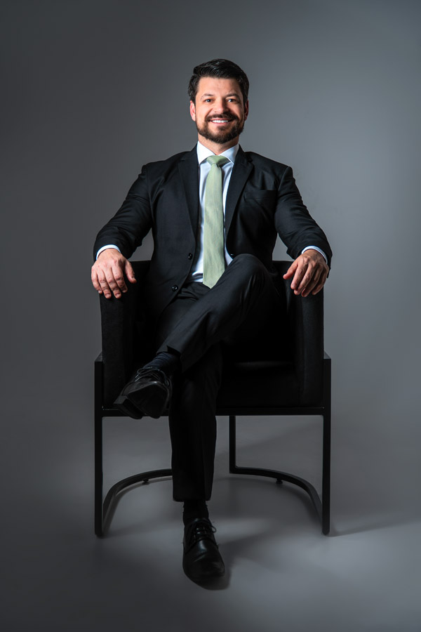 Homem de terno sorrindo amplamente com expressão confiante, corpo levemente inclinado para frente, com fundo escuro. Transmite carisma e abertura.