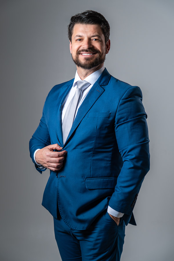 Homem com sorriso sutil, braços cruzados sobre a mesa, olhando diretamente para a câmera, com fundo escuro. Passa credibilidade e receptividade.