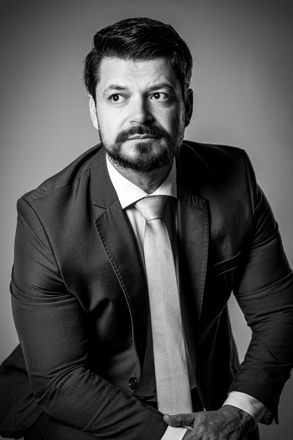 Homem de terno com olhar firme voltado para o lado, em fundo escuro, com expressão de foco e determinação. Imagem com contraste intenso.