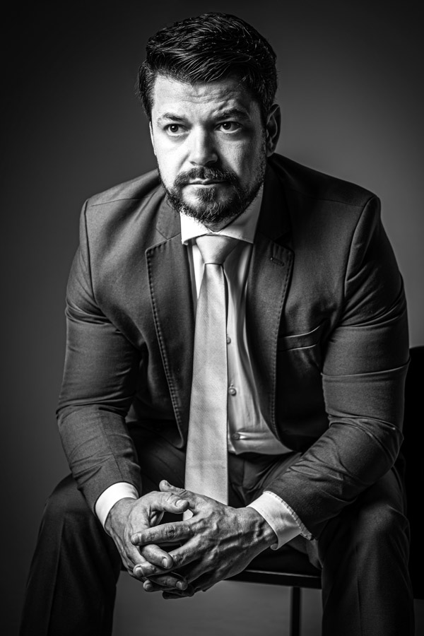 Homem sentado com perna cruzada e expressão neutra, vestindo terno, com fundo escuro. Posição relaxada que transmite profissionalismo.