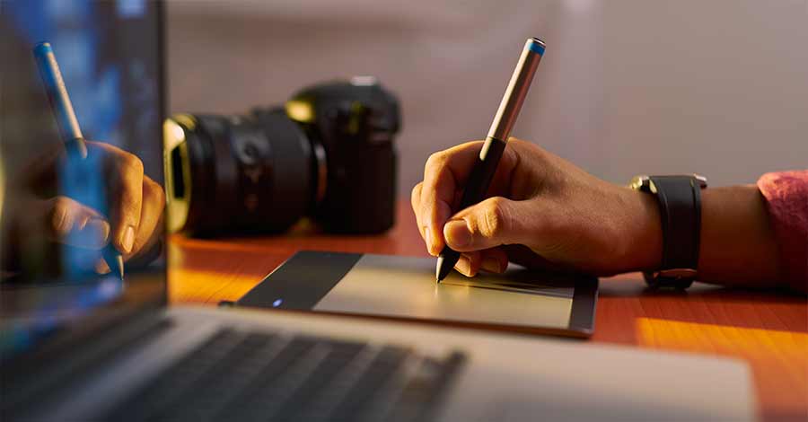 Mão segurando caneta digital e desenhando em mesa digitalizadora, com câmera DSLR desfocada ao fundo e notebook aberto.