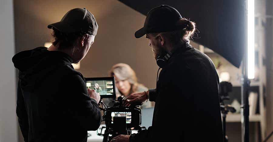 Dois cinegrafistas ajustando uma câmera profissional durante uma gravação em estúdio, com uma mulher desfocada ao fundo sendo filmada.