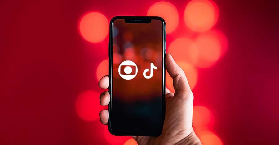 Mão segurando um smartphone com as logos da TV Globo e TikTok na tela, com fundo desfocado em tons de vermelho e luzes circulares.