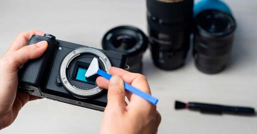 Pessoa limpando o sensor de uma câmera digital com uma ferramenta de limpeza específica, com lentes fotográficas desfocadas ao fundo