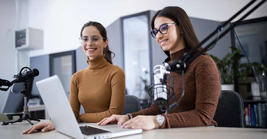 Duas mulheres sorridentes gravando um podcast em estúdio, com microfones profissionais e laptop sobre a mesa