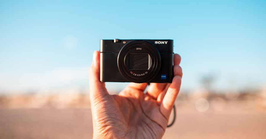 Mão segurando uma câmera compacta Sony apontada para a frente em ambiente externo iluminado.