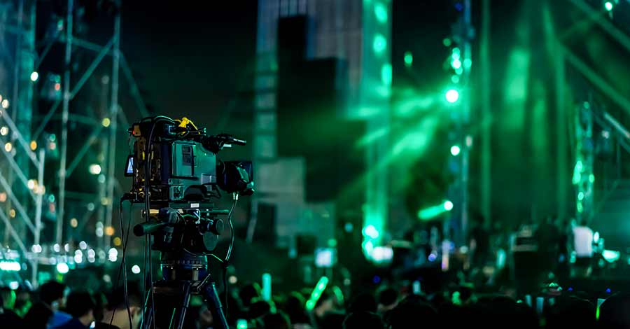 Câmera profissional montada em tripé filmando um show ou evento ao vivo, com iluminação verde intensa e público ao fundo.