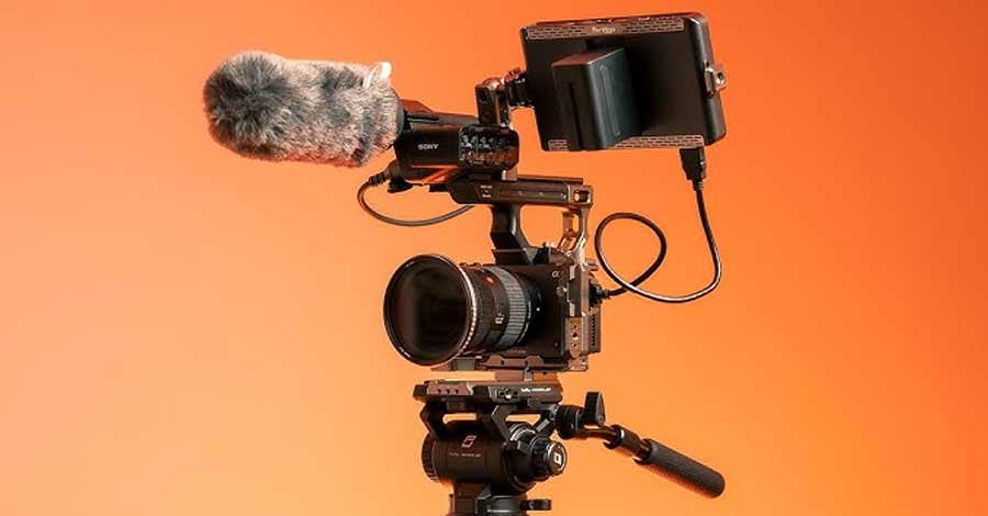 Câmera profissional equipada com microfone, monitor externo e rig completo sobre fundo laranja.