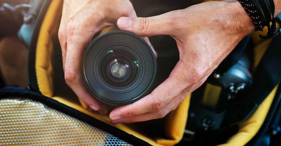 Mãos segurando uma lente de câmera fotográfica sobre uma mochila acolchoada de equipamentos.