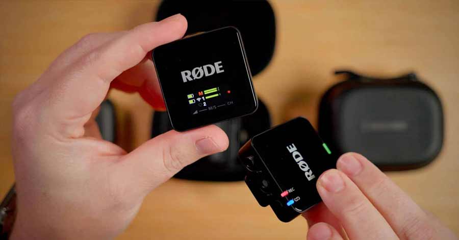 Duas unidades do microfone sem fio RØDE Wireless GO II sendo seguradas nas mãos, com visor mostrando níveis de áudio e estojo de carregamento ao fundo.