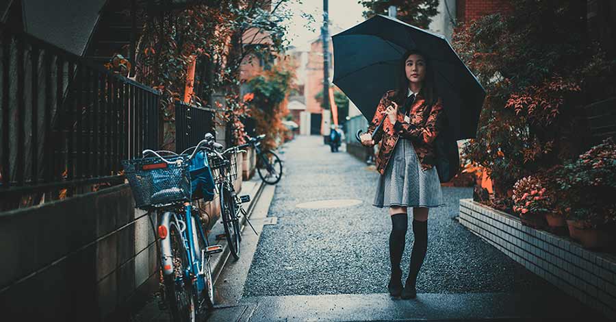 Mulher caminhando por uma rua estreita em dia chuvoso, segurando um guarda-chuva preto, com bicicletas estacionadas ao lado e plantas ao redor.