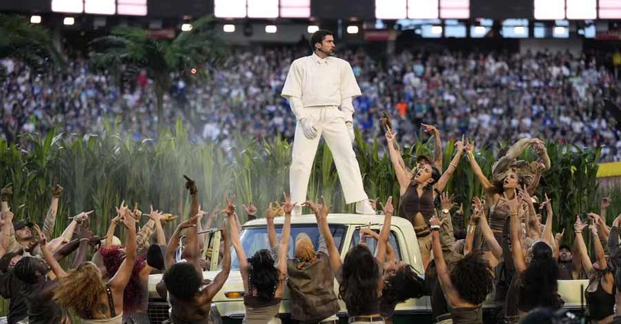 Artista vestido de branco sobre um carro cercado por dançarinos em coreografia coletiva diante de uma multidão em estádio.