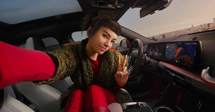 Mulher soprando bolhas de sabão ao lado de um carro BMW X2 azul estacionado ao ar livre, com paisagem ao fundo.