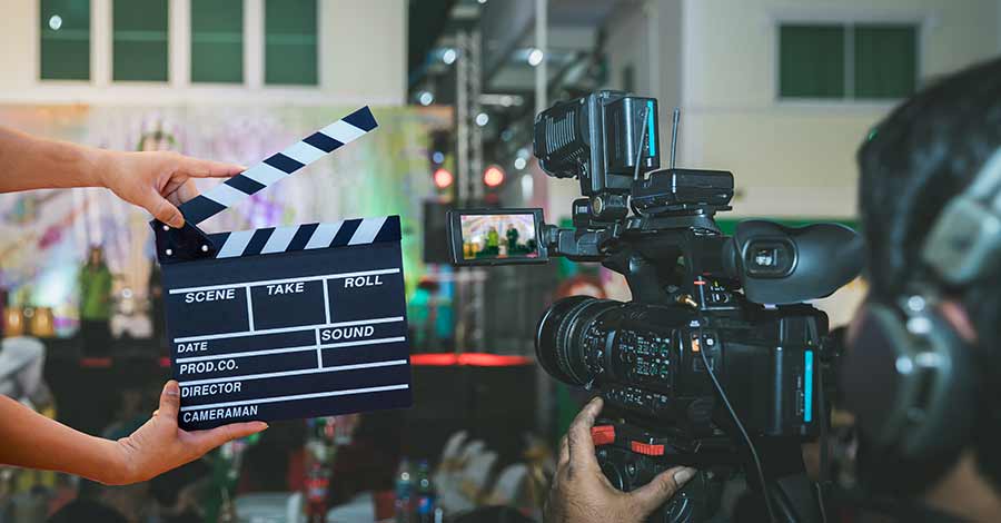 Claquete de cinema sendo segurada em frente a uma câmera profissional durante uma gravação em ambiente interno.