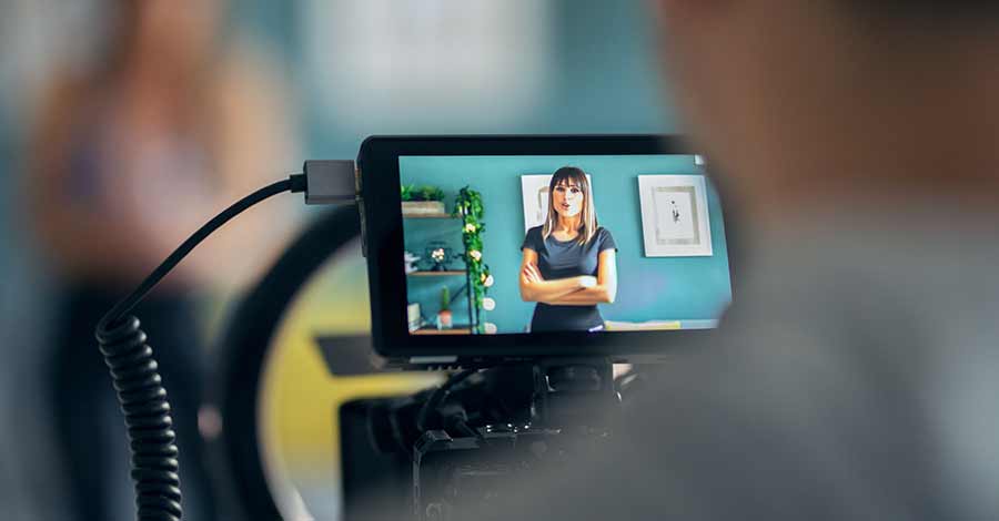 Mulher sendo gravada em vídeo dentro de um estúdio, aparecendo no monitor de uma câmera enquanto fala para a gravação.
