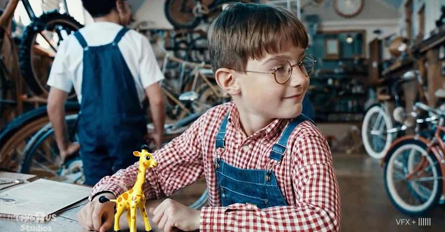 Menino com óculos e avental sentado em oficina de bicicletas, brincando com um pequeno brinquedo de girafa sobre a mesa.